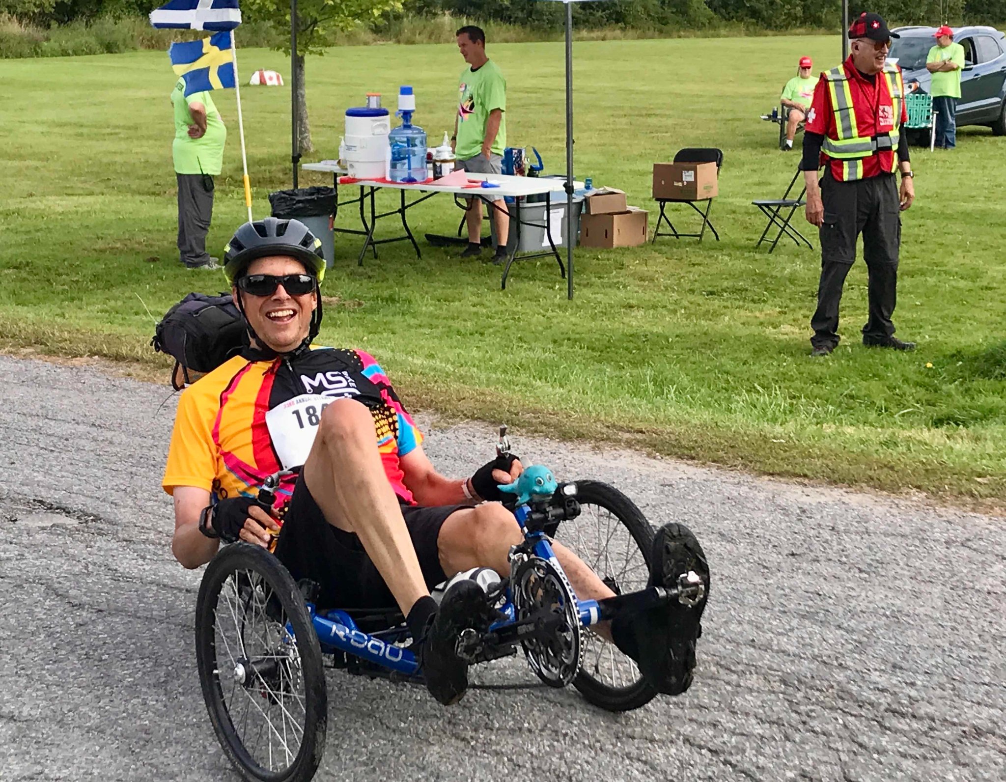 Photo of cyclist on recumbent tricycle on the road in front of a support tent, smiling at the camera