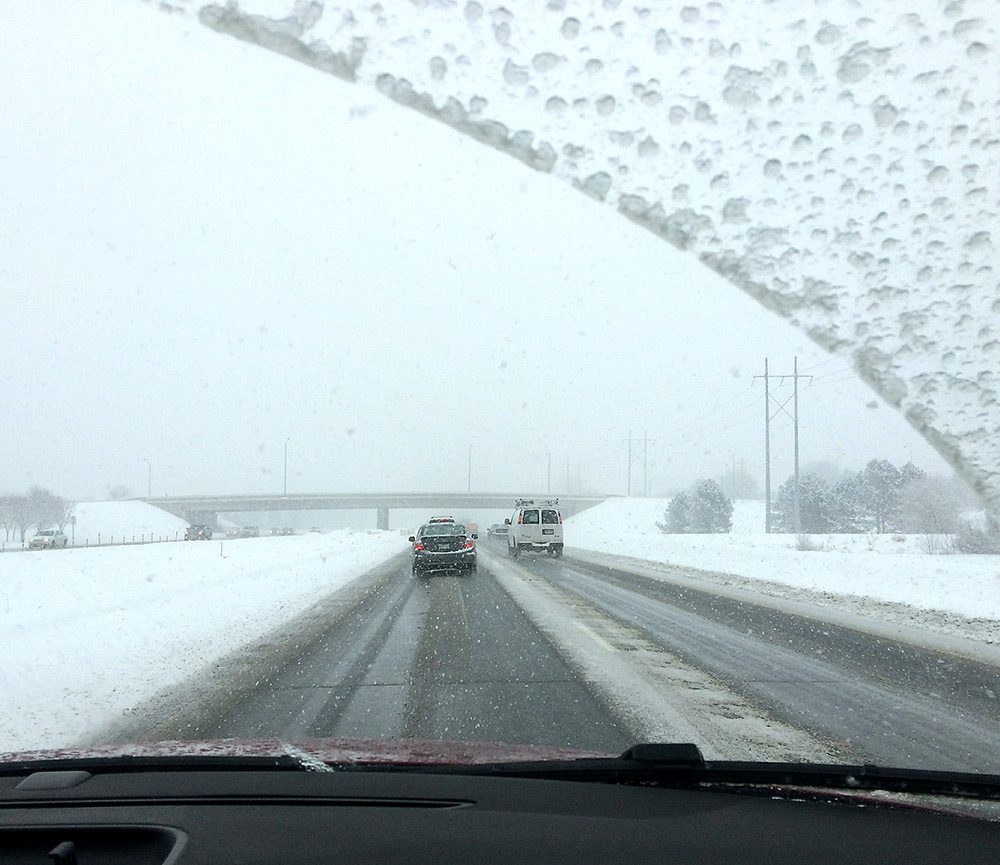 View from the front window of a car, on a grey day, while driving on a highway. It is snowing, the medians are snow-covered, and there is some ice on the windshield.