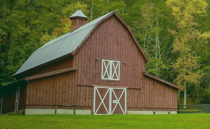 A burnt sienna coloured barn with white trim doors in front of a treed area.