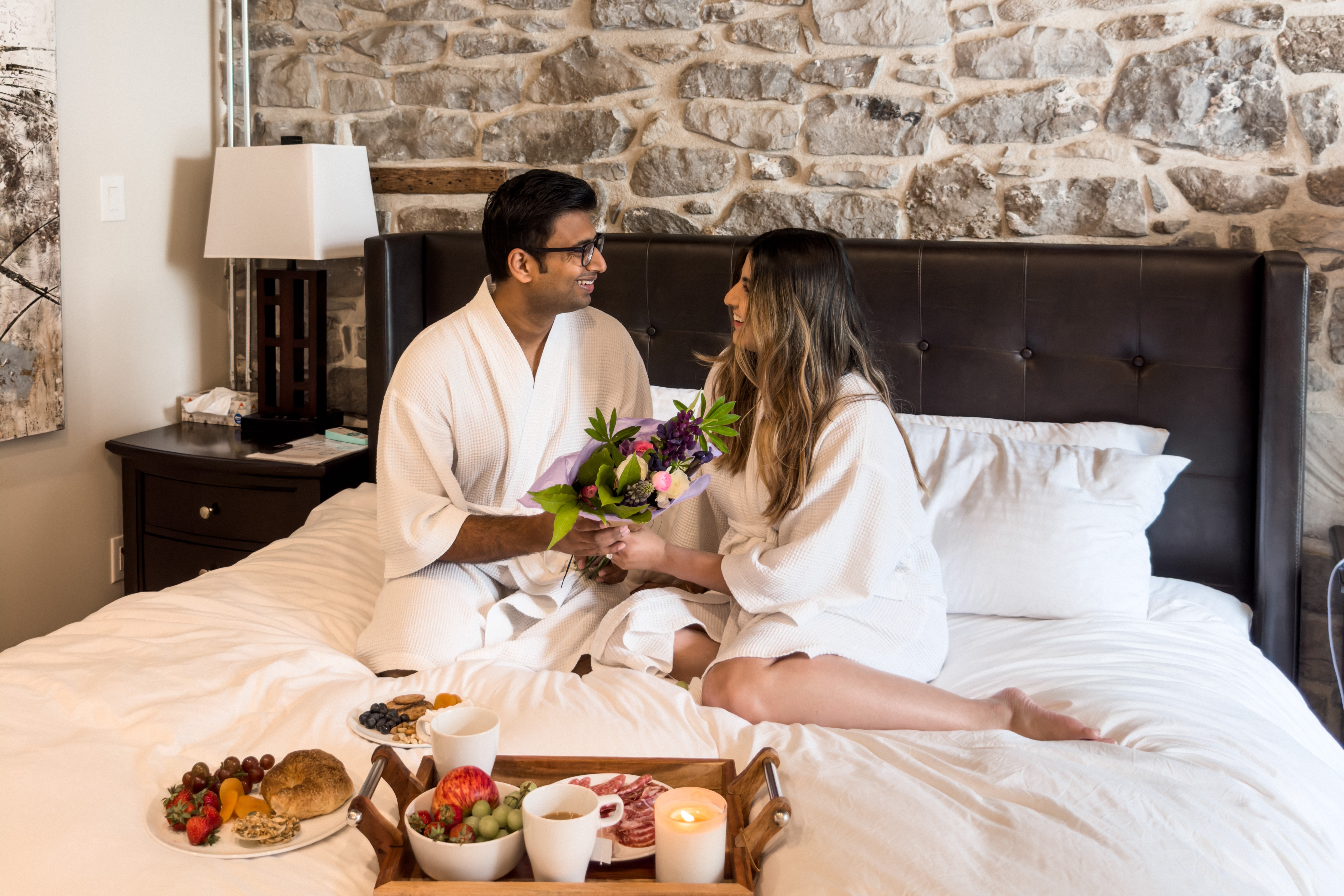 Couple in robes sit affectionately on bed. The two are holding flowers from Wilding Acres. There is a breakfast platter placed on the bed.
